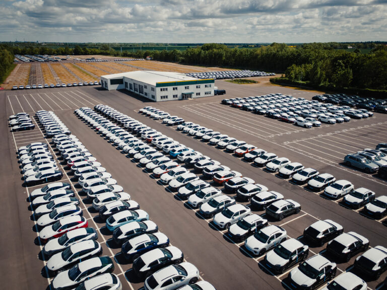 Luftaufnahme eines großen Parkplatzes mit Reihen von weißen Autos, die ein zentrales Gebäude umgeben, mit grünen Bäumen und Feldern im Hintergrund unter einem bewölkten Himmel.