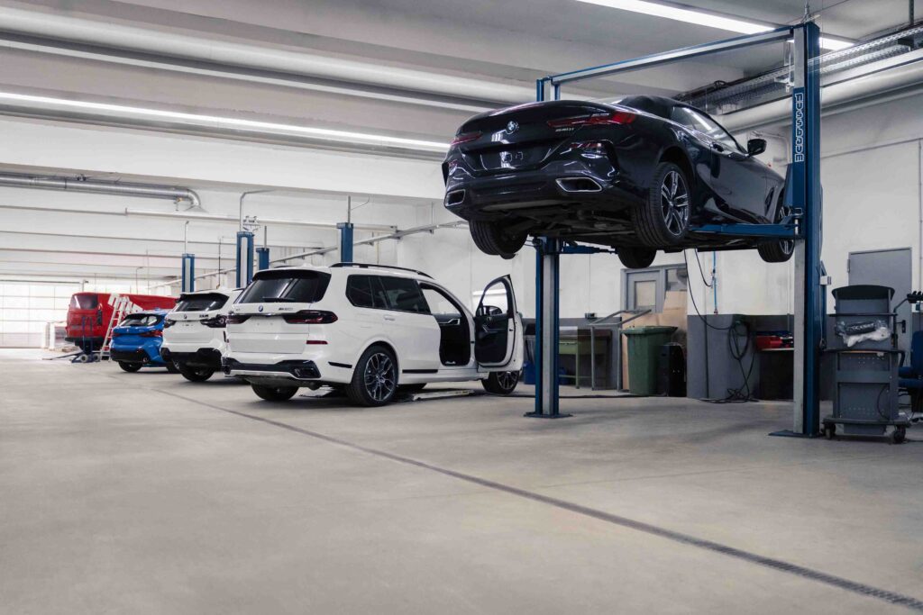 A spacious, modern automotive workshop with several cars; one black car is elevated on a hydraulic lift, while white and blue vehicles are parked nearby. Various tools and equipment are visible around the workshop.
