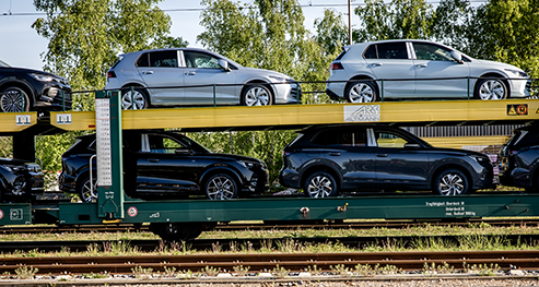 Mehrere Neuwagen werden für den Transport auf einen zweistöckigen Waggon verladen, im Hintergrund sind Bäume und Gleise zu sehen.