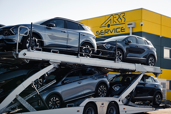 Ein mit mehreren Geländewagen beladener Autotransportanhänger ist vor einem gelb-grauen Gebäude mit der Aufschrift ARS SERVICE an der Fassade geparkt.