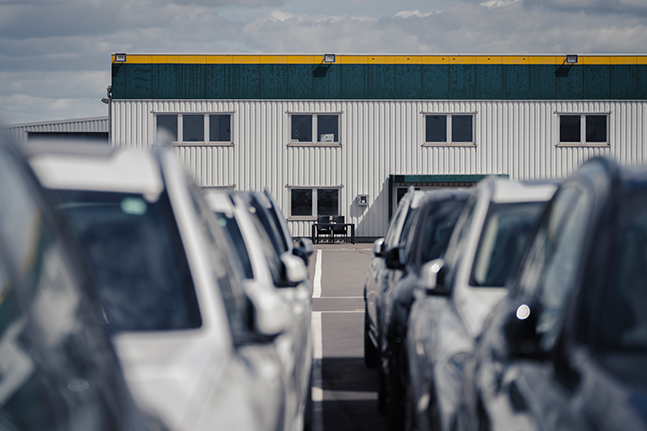 Reihen geparkter weißer Fahrzeuge säumen beide Seiten eines Parkplatzes und führen zu einem grauen Industriegebäude mit gelber Fassade unter einem bewölkten Himmel.