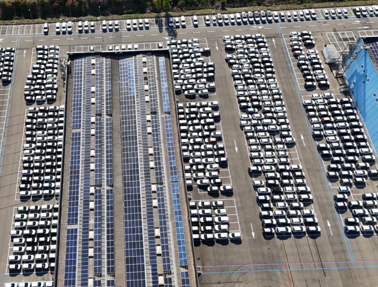 Luftaufnahme eines großen Parkplatzes mit Reihen von weißen Autos. Ein langer Abschnitt in der Mitte ist mit Sonnenkollektoren bedeckt. Die Szene ist organisiert und geordnet, mit klaren Parklinien sichtbar.
