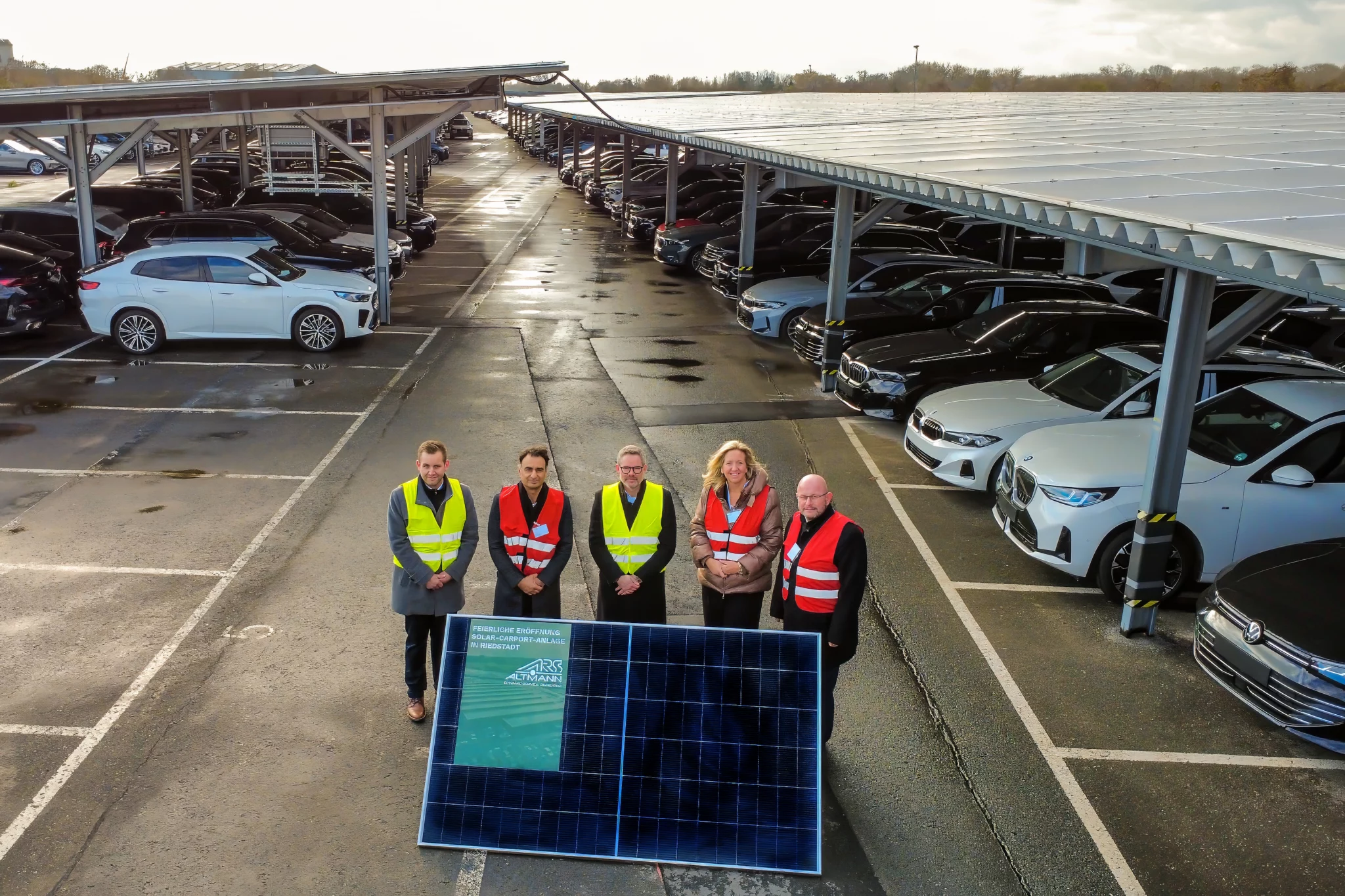 Fünf Personen in Sicherheitswesten stehen vor einem Solarmodul und posieren auf einem großen Parkplatz mit vielen Autos, die an einem bewölkten Tag unter Solarmodulüberdachungen geparkt sind.